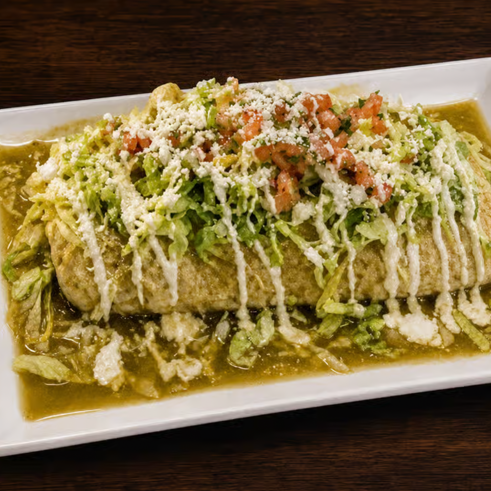 Enchiladas topped with shredded lettuce, diced tomatoes, crumbled cheese, and cream, served in green sauce on a white platter.