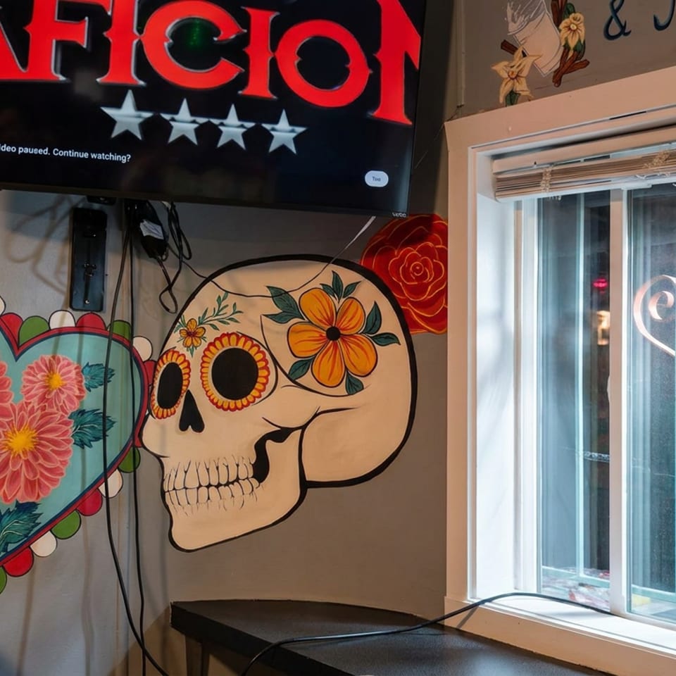 Colorful wall murals depict a flower-filled cactus heart and decorated skull beside a television and bright restaurant window.