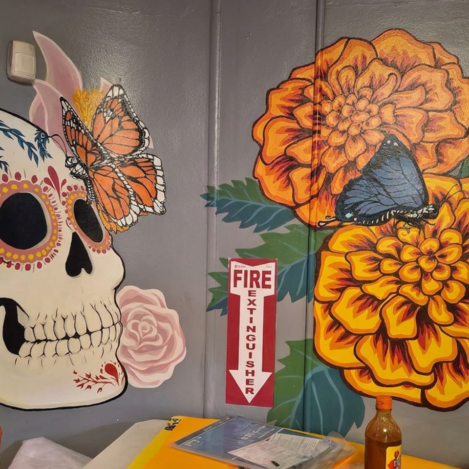 Colorful mural features a floral sugar skull with butterflies beside large orange flowers and a fire extinguisher sign.
