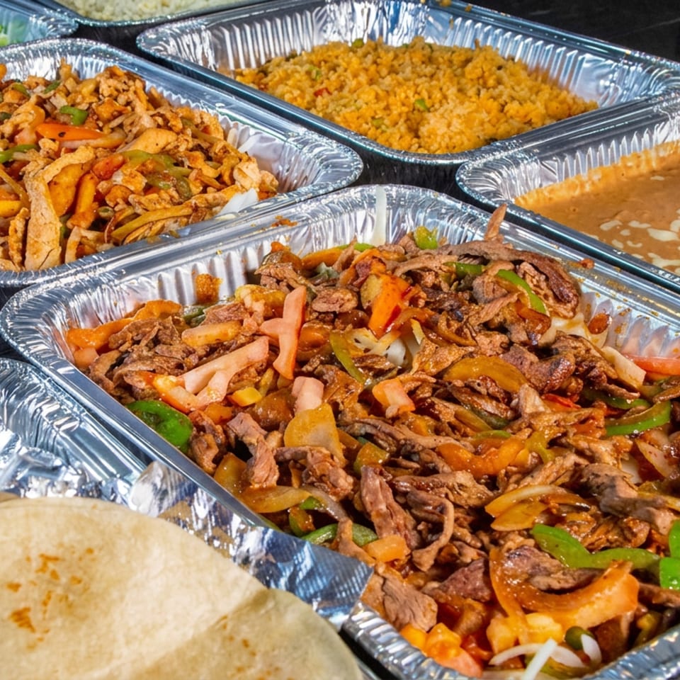 Aluminum trays display fajita-style meats and vegetables, Mexican rice, beans, salad, and tortillas on a restaurant counter.