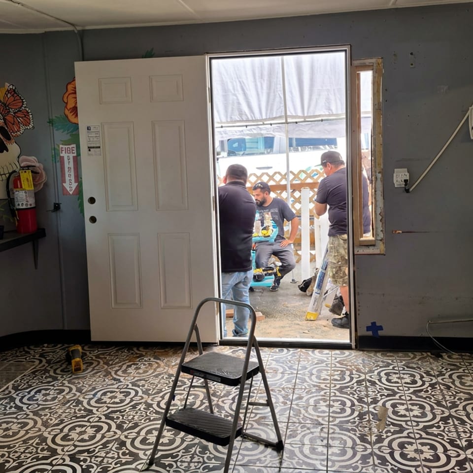 Restaurant interior with patterned tile floor, colorful skull mural, counter supplies, and people gathered outside the open door.